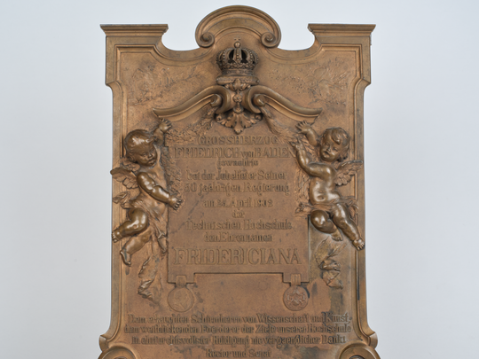 Gedenkplakette anlässlich der Verleihung des Beinamens Fridericiana an die Technische Hochschule Karlsruhe, Details am oberen Rand: Herrscherkrone (mittig), links daneben die Eule als Symbol der Wissenschaft, rechts neben der Krone Symbole für Abteilungen der Hochschule (schneckenförmige Dekoration für die Architektur, Glaskolben für die Chemie, Erdkugel für das Bauingenieur- und Vermessungswesen, gezackter Blitz für die Elektrotechnik, Zahnrad für den Maschinenbau, Nadelzweig für das Forstwesen). Fotografie: Amadeus Bramsiepe.