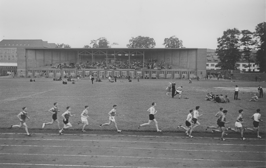 Tribüne des Hochschulstadions, Ansicht von Osten, im Vordergrund Sportler auf der Laufbahn, ca. 1950, Fotografie: Schlitz. KIT-Archiv 28010/I 3661.