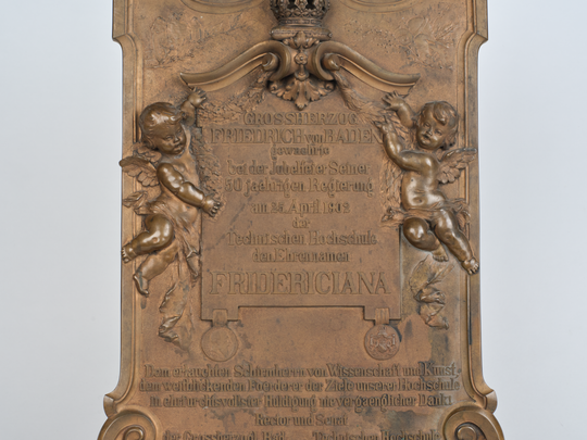 Gedenkplakette anlässlich der Verleihung des Beinamens Fridericiana an die Technische Hochschule Karlsruhe, Details in der Mitte: von geflügelten Kinderfiguren gehaltene Tafel mit Angabe des Widmungsanlasses, an dieser hängend Medaillons mit dem Portrait des Großherzogs Friedrich I. und dem badischen Landeswappen. Fotografie: Amadeus Bramsiepe.