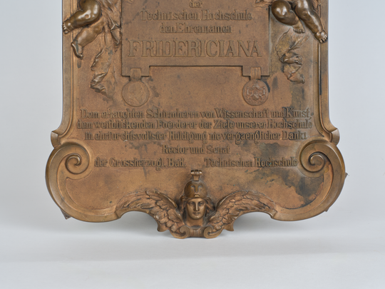 Gedenkplakette anlässlich der Verleihung des Beinamens Fridericiana an die Technische Hochschule Karlsruhe, Details unten: Dankadresse von Rektor und Senat an Großherzog Friedrich I. von Baden, darunter der Kopf von Pallas Athene auf Flügeln. Fotografie: Amadeus Bramsiepe.