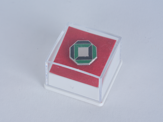 Anstecknadel mit dem Logo der Universität Karlsruhe, ca. 1990, 12,5 mm, Metall und Kunstharz. KIT-Archiv 28502/68. Fotografie: Amadeus Bramsiepe.